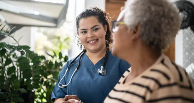 Home caregiver wearing a stethoscope around her neck helping senior woman walking at home